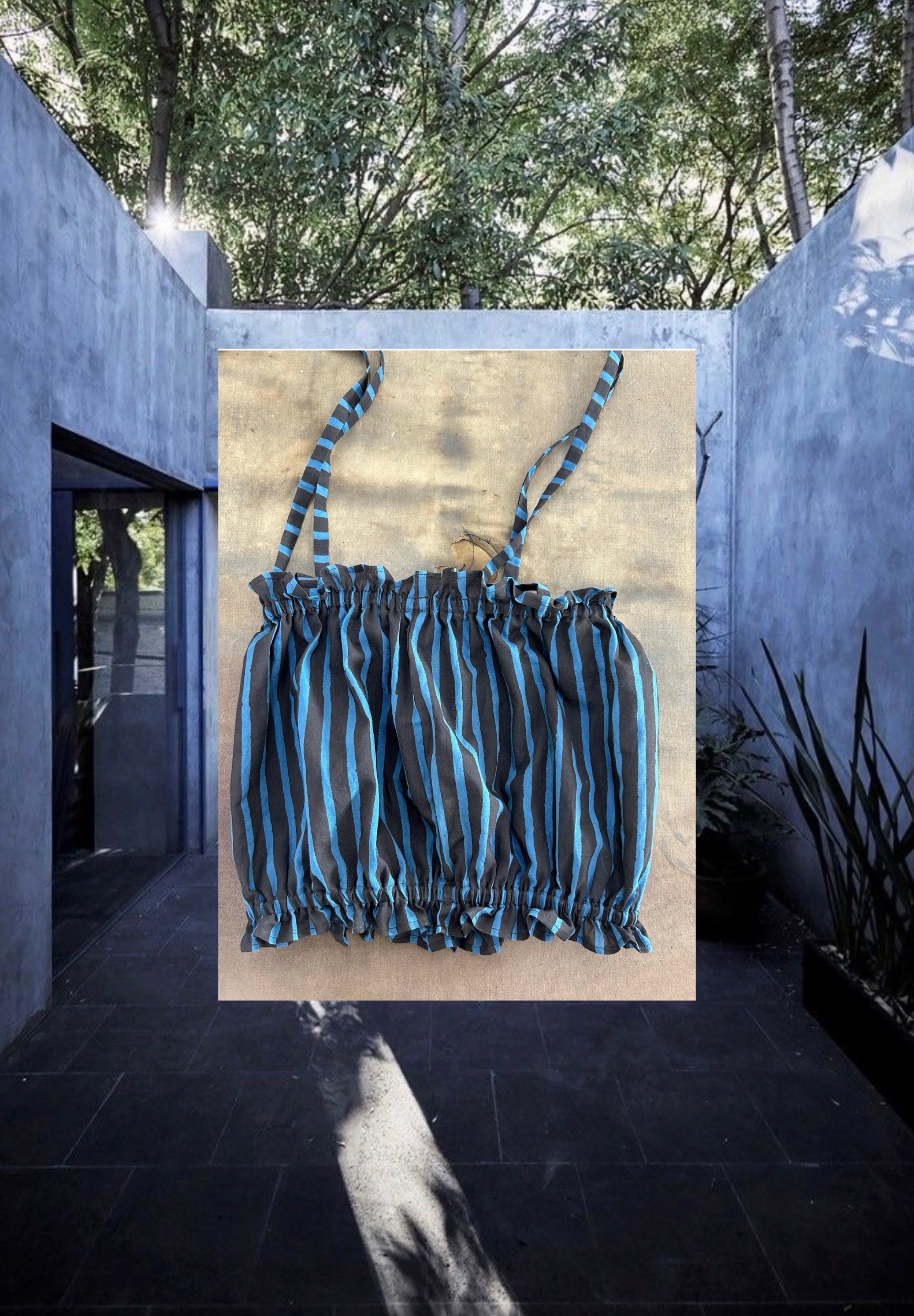 A sleeveless children's top with a ruched, gathered design and thin shoulder straps, featuring a black base with bright blue vertical stripes. The fabric has a lightweight, airy appearance, and the hem and neckline are elasticated for a snug fit.