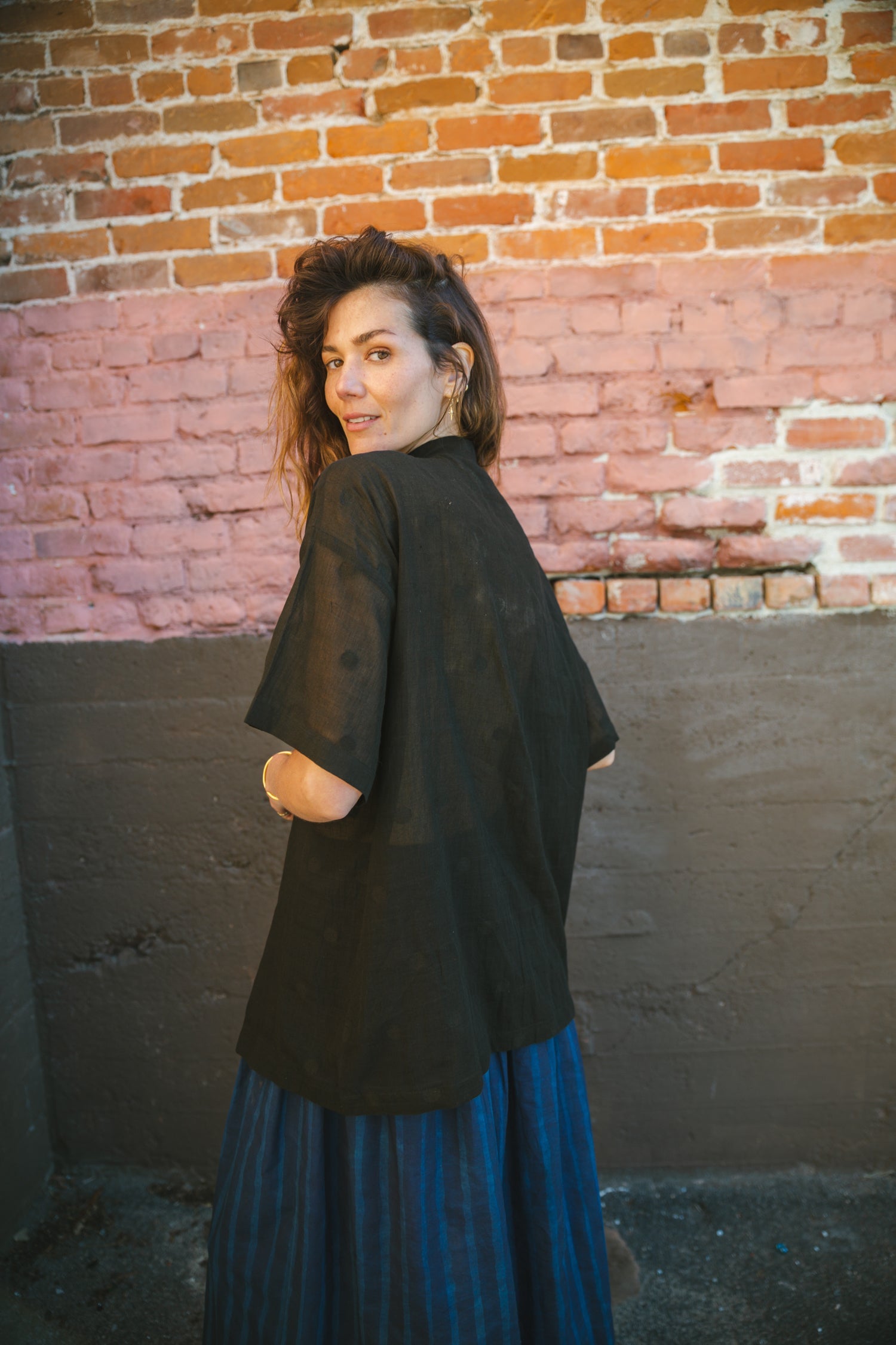 A black, short-sleeved button-up shirt made of a lightweight, semi-sheer fabric. It has a relaxed, oversized fit, a structured collar, and contrasting gold buttons running down the front. The breathable material and loose silhouette give it an effortlessly casual yet polished look.