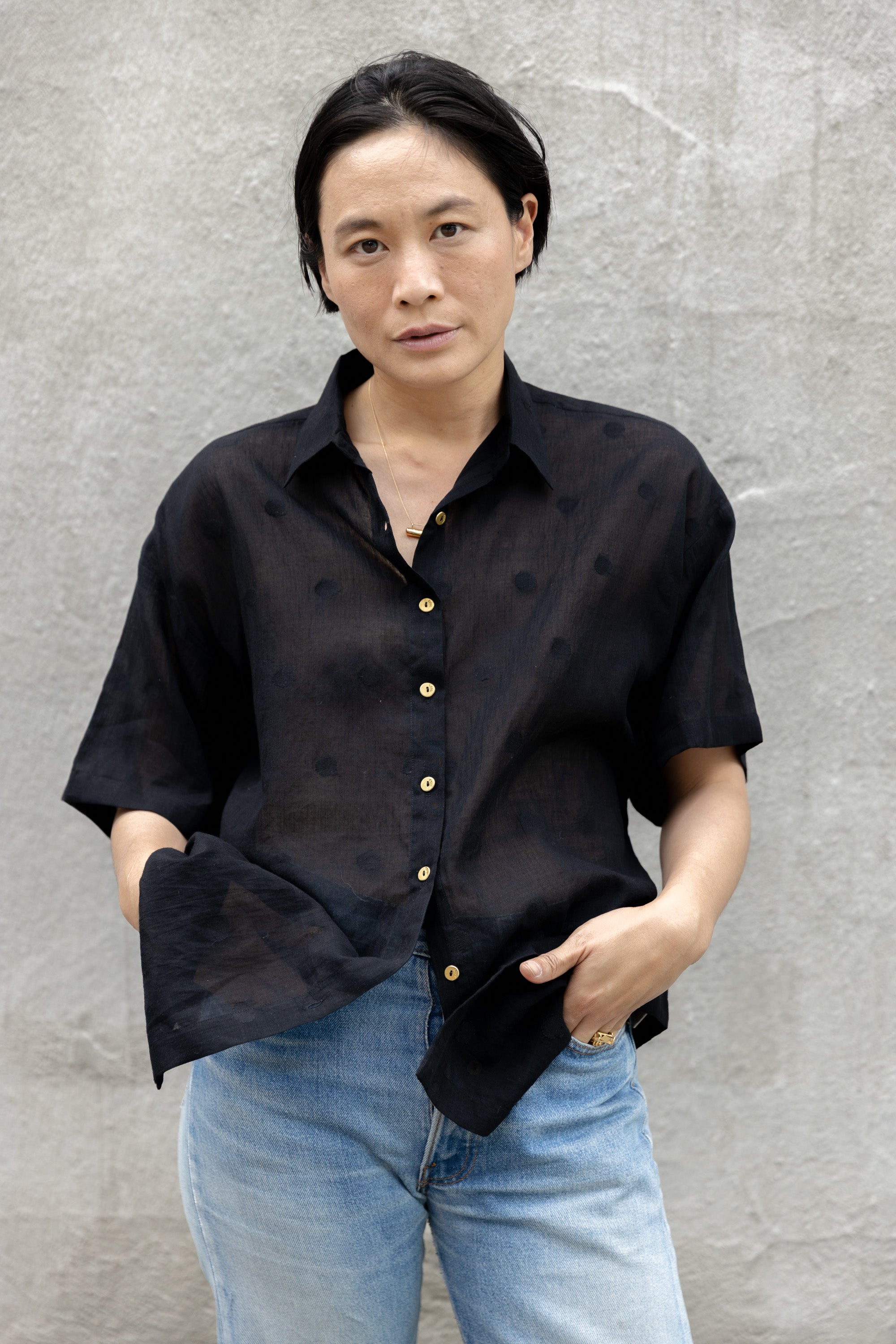 A black, short-sleeved button-up shirt made of a lightweight, semi-sheer fabric. It has a relaxed, oversized fit, a structured collar, and contrasting gold buttons running down the front. The breathable material and loose silhouette give it an effortlessly casual yet polished look.
