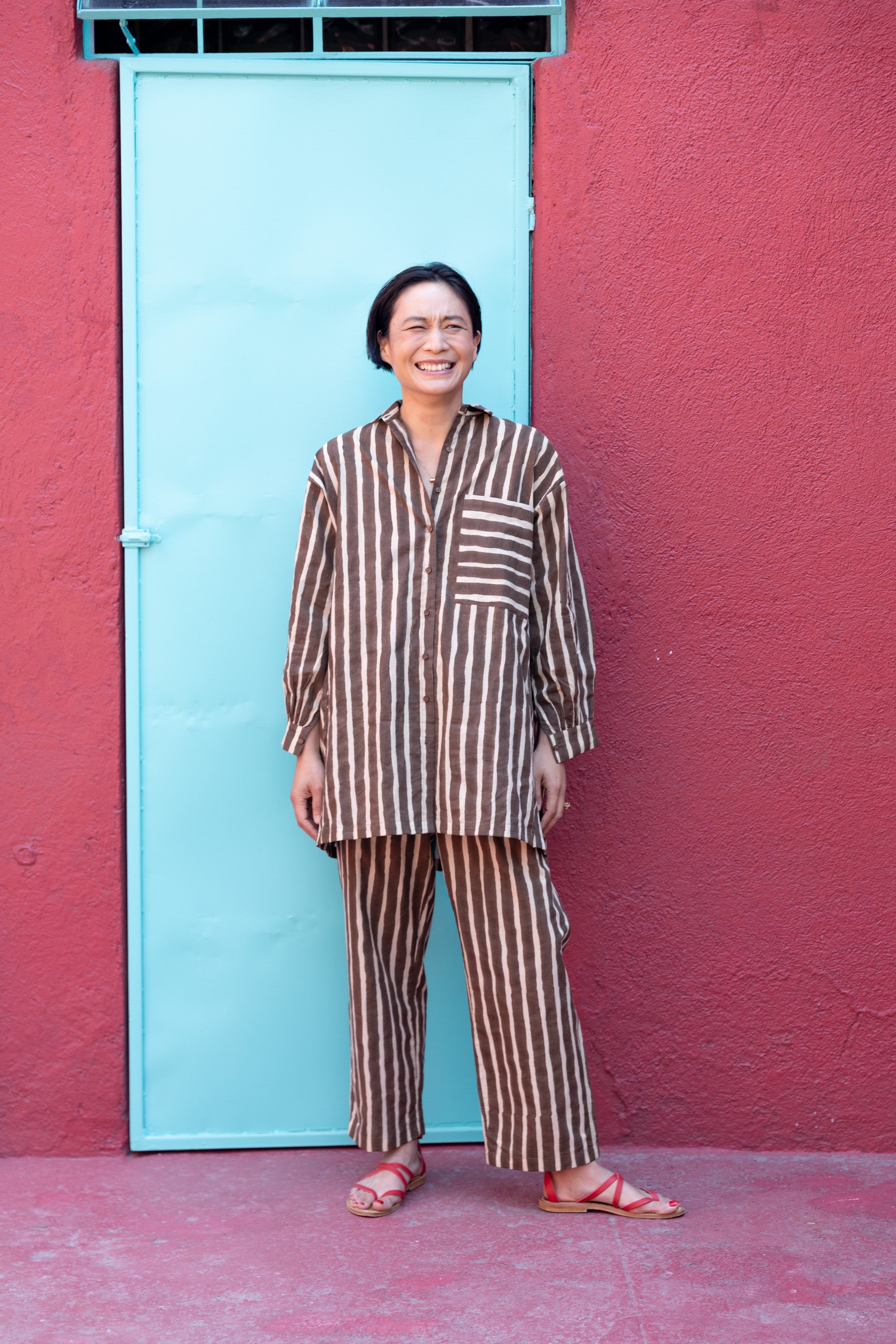A brown and white striped long-sleeve shirt with a relaxed, oversized fit. The shirt features a classic pointed collar and a V-neck opening without buttons, giving it an effortless and casual look. A single chest pocket with horizontal stripes contrasts with the vertical pattern on the rest of the shirt, adding visual interest. The lightweight fabric drapes comfortably, making it ideal for a relaxed yet stylish outfit.