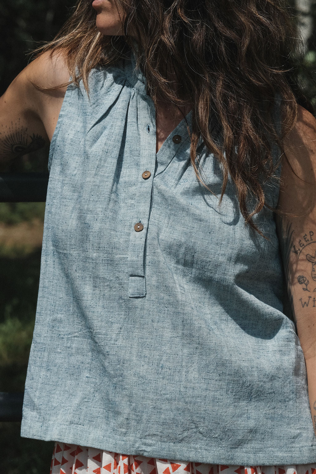 Sleeveless chambray blue top made from soft, handwoven organic cotton. Features a short button placket at the front and a subtle pleat detail below the collar, creating a clean and structured look. The stand collar adds a modern touch, while the breathable, lightweight fabric ensures comfort in warmer weather. Naturally dyed and ethically crafted, this top is perfect for effortless, everyday wear.