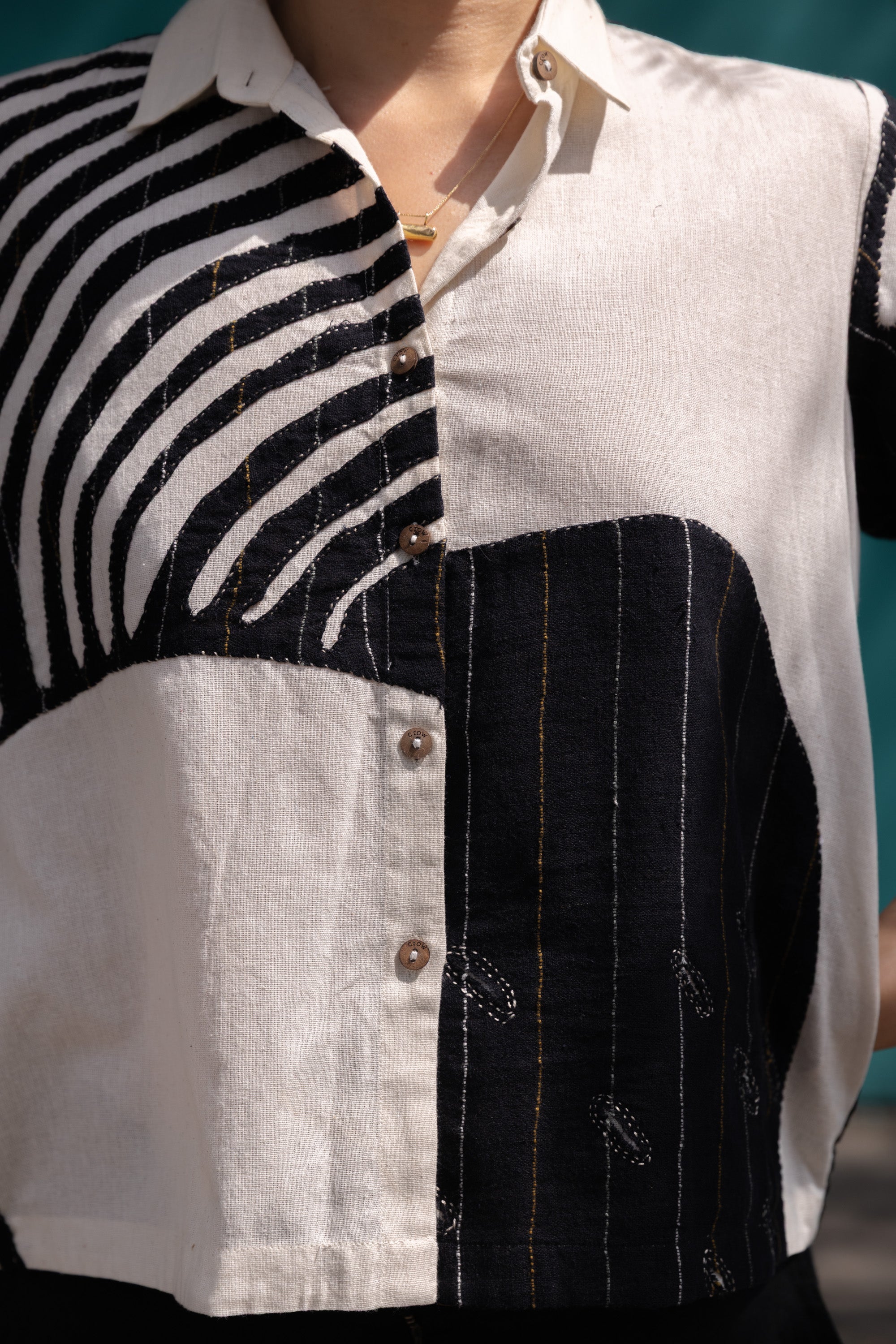 A short-sleeved, button-up shirt featuring a striking asymmetrical patchwork design in black, white, and beige. The fabric has a lightweight, textured feel, crafted from breathable natural fibers. The pattern blends bold geometric lines, organic curves, and subtle embroidered details, creating a dynamic contrast. The classic pointed collar adds a structured touch, while the relaxed, boxy fit gives it an effortlessly modern, artistic appeal.