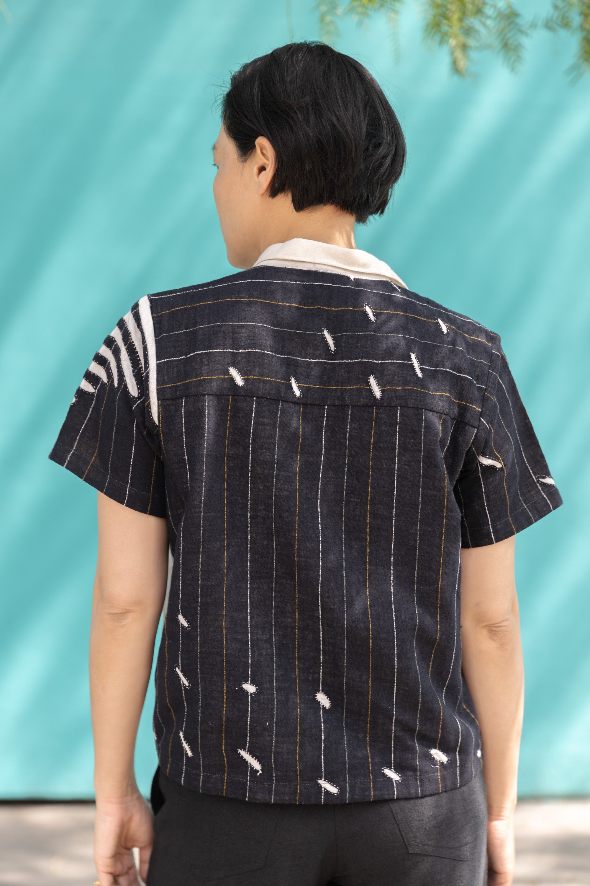 A short-sleeved, button-up shirt featuring a striking asymmetrical patchwork design in black, white, and beige. The fabric has a lightweight, textured feel, crafted from breathable natural fibers. The pattern blends bold geometric lines, organic curves, and subtle embroidered details, creating a dynamic contrast. The classic pointed collar adds a structured touch, while the relaxed, boxy fit gives it an effortlessly modern, artistic appeal.