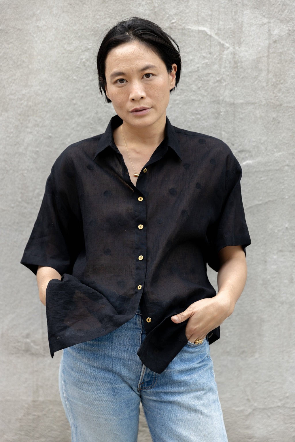 A black, short-sleeved button-up shirt made of a lightweight, semi-sheer fabric. It has a relaxed, oversized fit, a structured collar, and contrasting gold buttons running down the front. The breathable material and loose silhouette give it an effortlessly casual yet polished look.