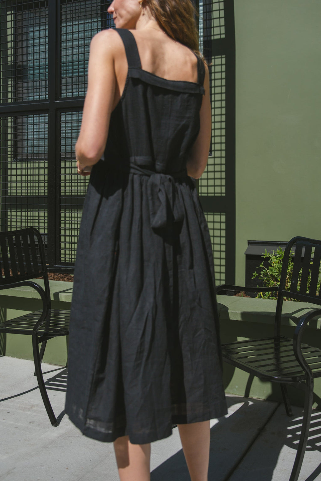 This is a black sleeveless midi dress with a structured square neckline and wide shoulder straps. The dress features a loose, flowing silhouette with a gathered waistline that adds volume and movement. Crafted from a lightweight, breathable fabric, it drapes effortlessly, making it perfect for warm weather. Functional side pockets enhance its practicality, while the relaxed fit exudes a laid-back yet refined charm. The minimalist design and deep black hue create a versatile piece that can be styled casually