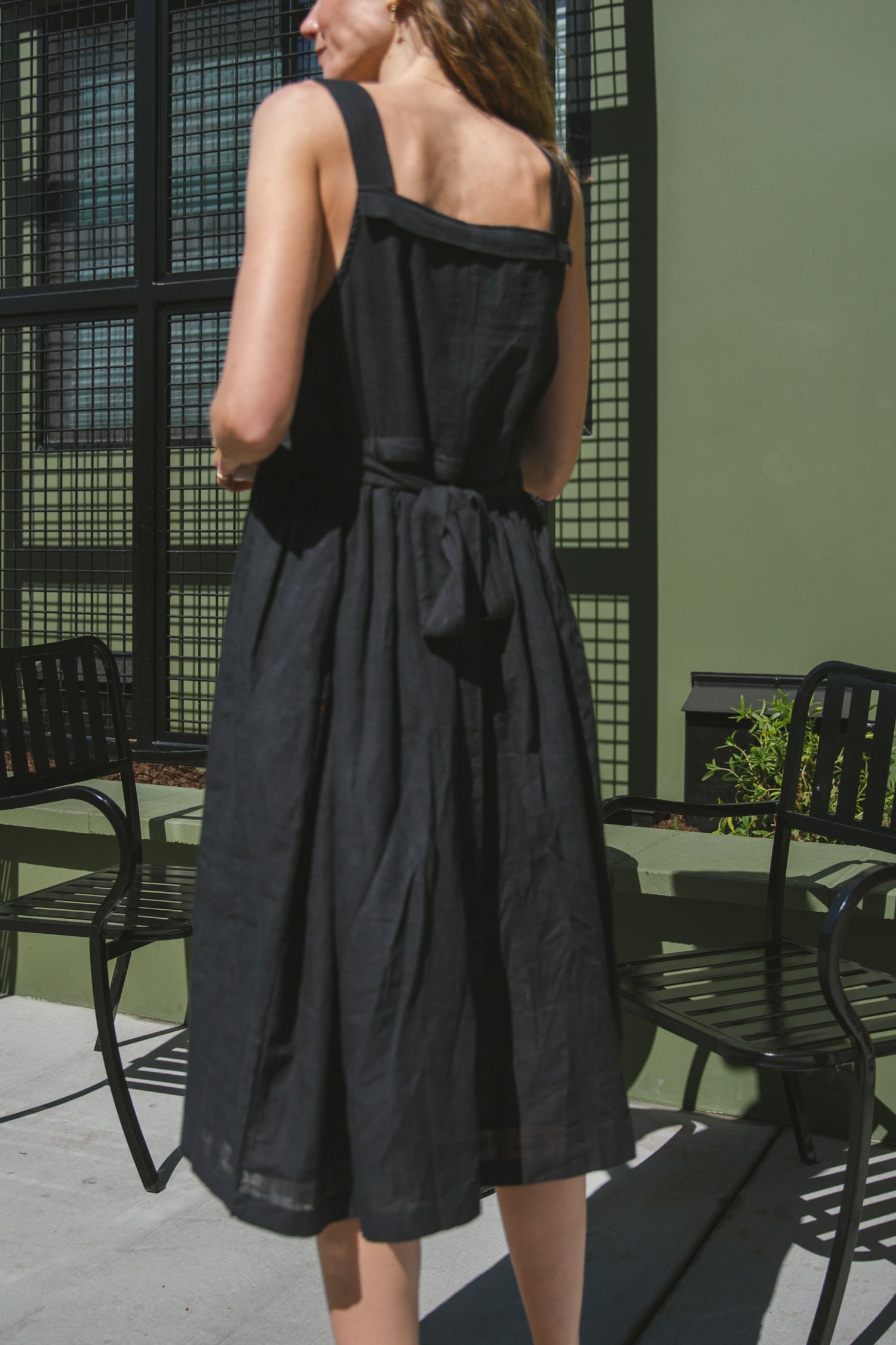 This is a black sleeveless midi dress with a structured square neckline and wide shoulder straps. The dress features a loose, flowing silhouette with a gathered waistline that adds volume and movement. Crafted from a lightweight, breathable fabric, it drapes effortlessly, making it perfect for warm weather. Functional side pockets enhance its practicality, while the relaxed fit exudes a laid-back yet refined charm. The minimalist design and deep black hue create a versatile piece that can be styled casually