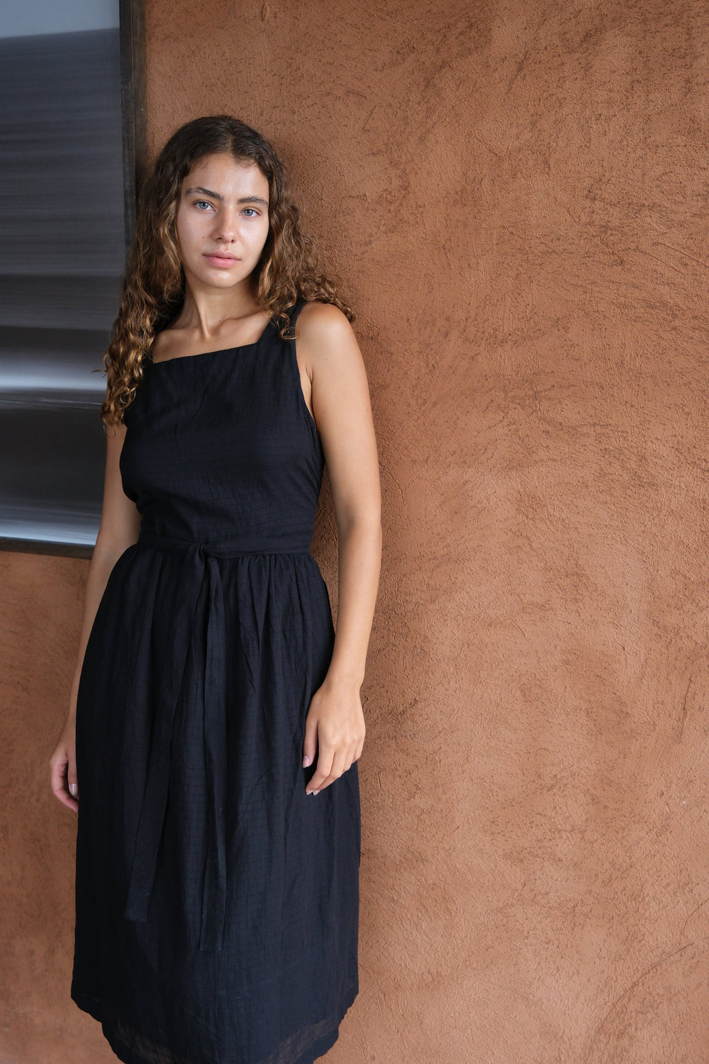 Introducing The Classic Hitch Dress, a collaboration between Audrey E Leary and World of Crow. This black midi-length dress features a square neck and a wrap-on design with a drawstring at the waist. Sleeveless with a straight hem, it is crafted from soft, lightweight fabric that offers a comfortable fit over the bust and a relaxed fit around the body. True to size, this dress combines elegance with ease for any occasion.