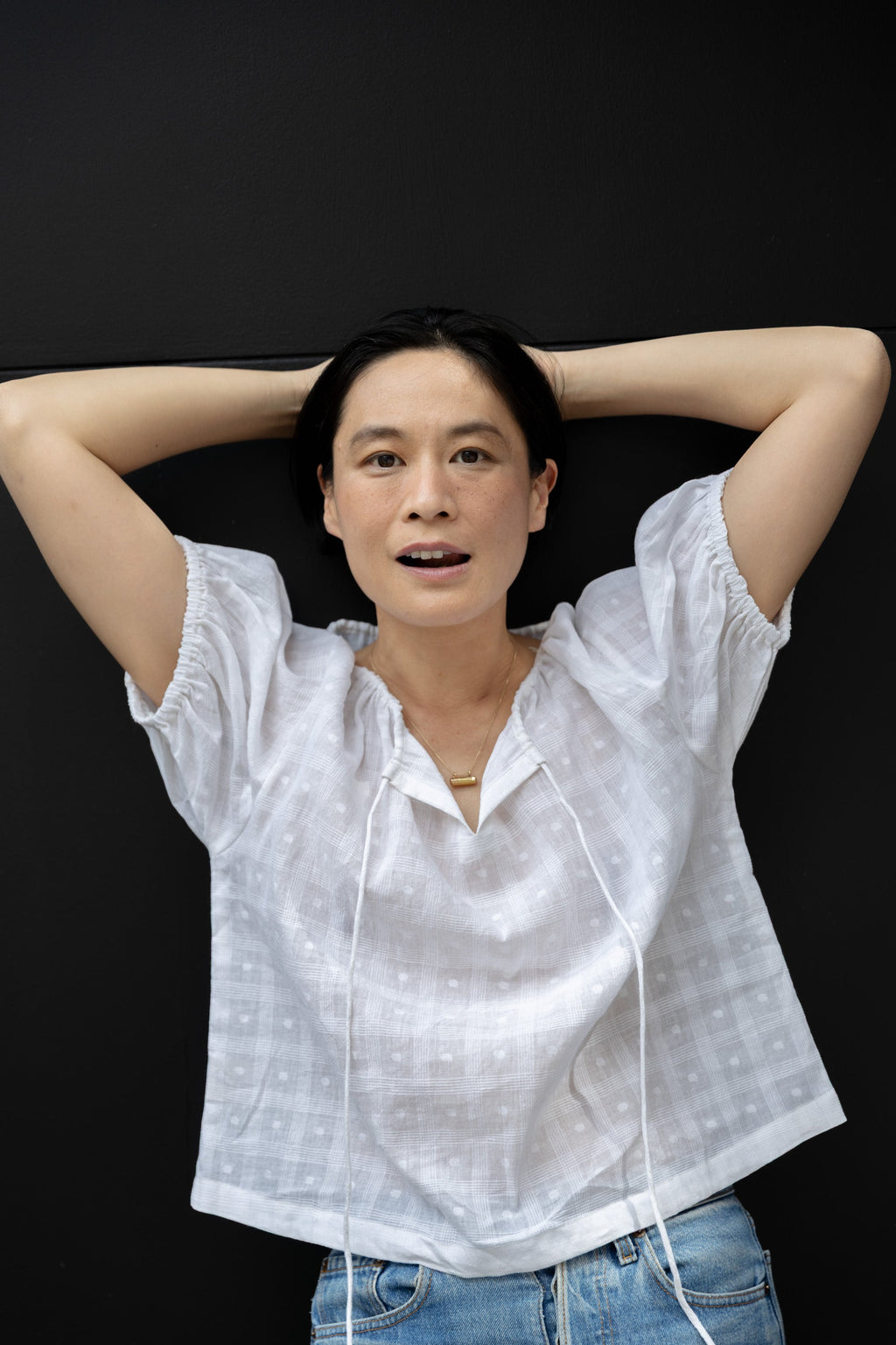 Lightweight white cotton top with a subtle textured weave, featuring a relaxed fit and airy design. The blouse has a gathered scoop neckline with delicate tie details, adding a touch of softness. Puff sleeves with gentle pleats create volume, while the slightly cropped hem gives it a modern, effortless feel.