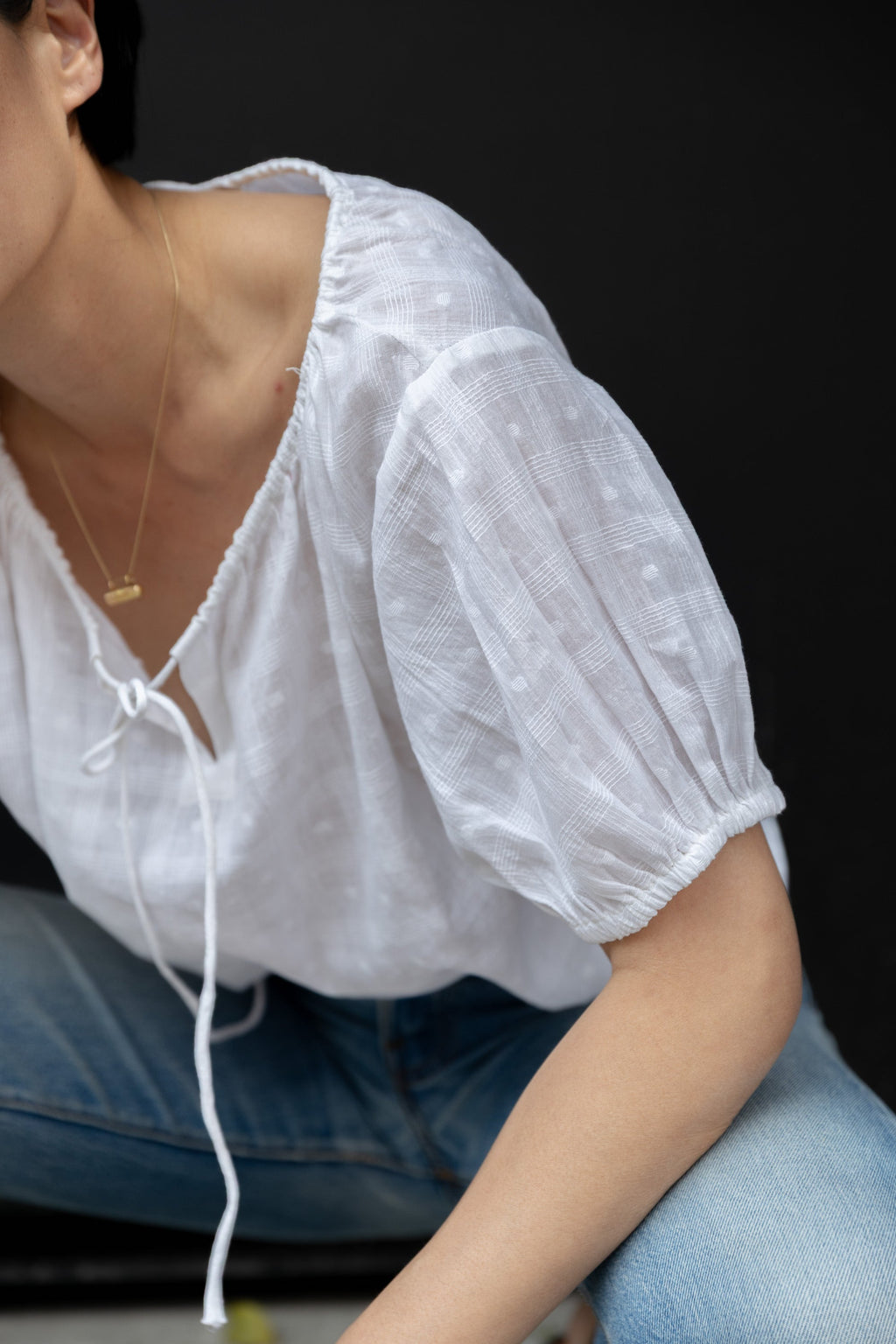 Lightweight white cotton top with a subtle textured weave, featuring a relaxed fit and airy design. The blouse has a gathered scoop neckline with delicate tie details, adding a touch of softness. Puff sleeves with gentle pleats create volume, while the slightly cropped hem gives it a modern, effortless feel.