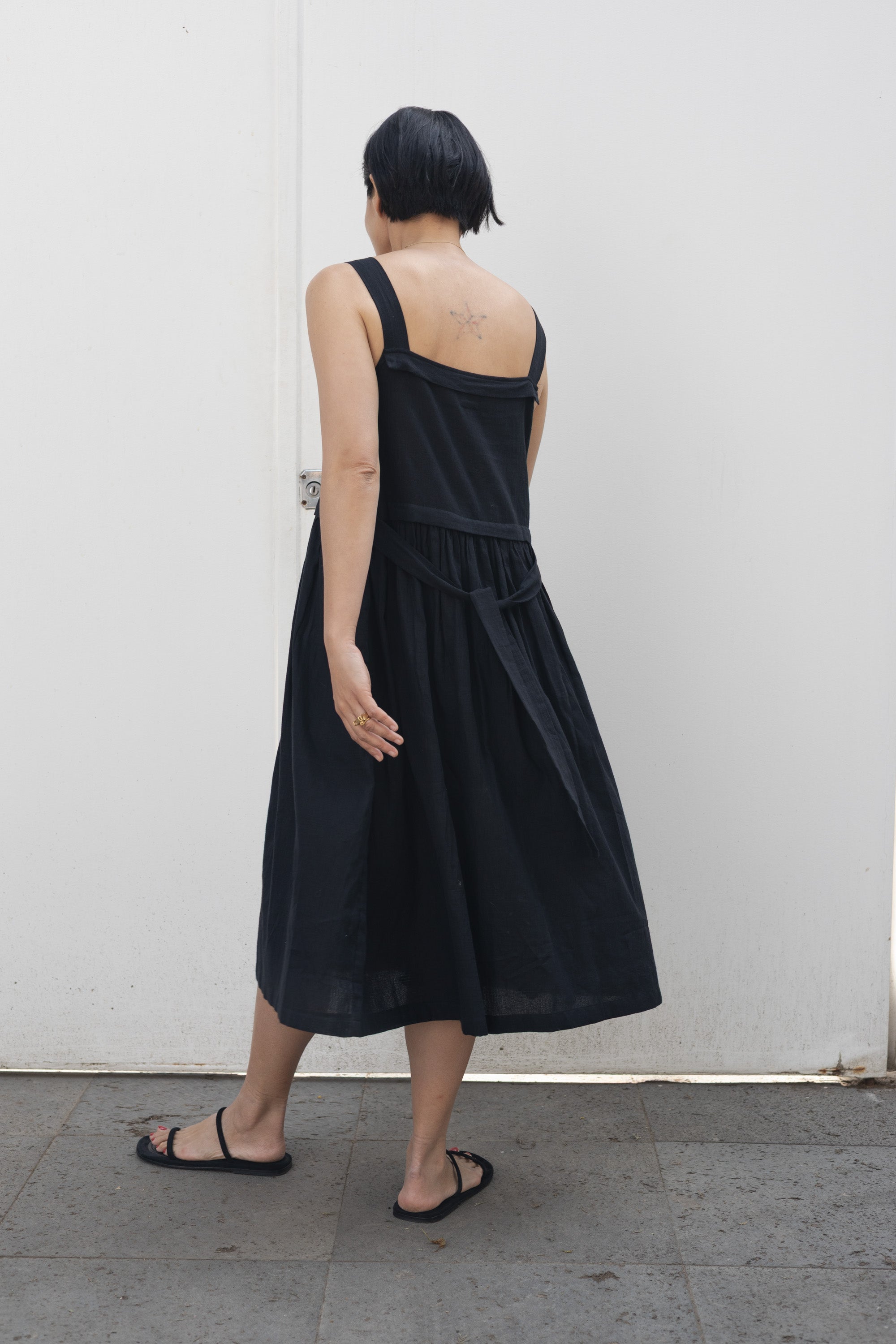 This is a black sleeveless midi dress with a structured square neckline and wide shoulder straps. The dress features a loose, flowing silhouette with a gathered waistline that adds volume and movement. Crafted from a lightweight, breathable fabric, it drapes effortlessly, making it perfect for warm weather. Functional side pockets enhance its practicality, while the relaxed fit exudes a laid-back yet refined charm. The minimalist design and deep black hue create a versatile piece that can be styled casually