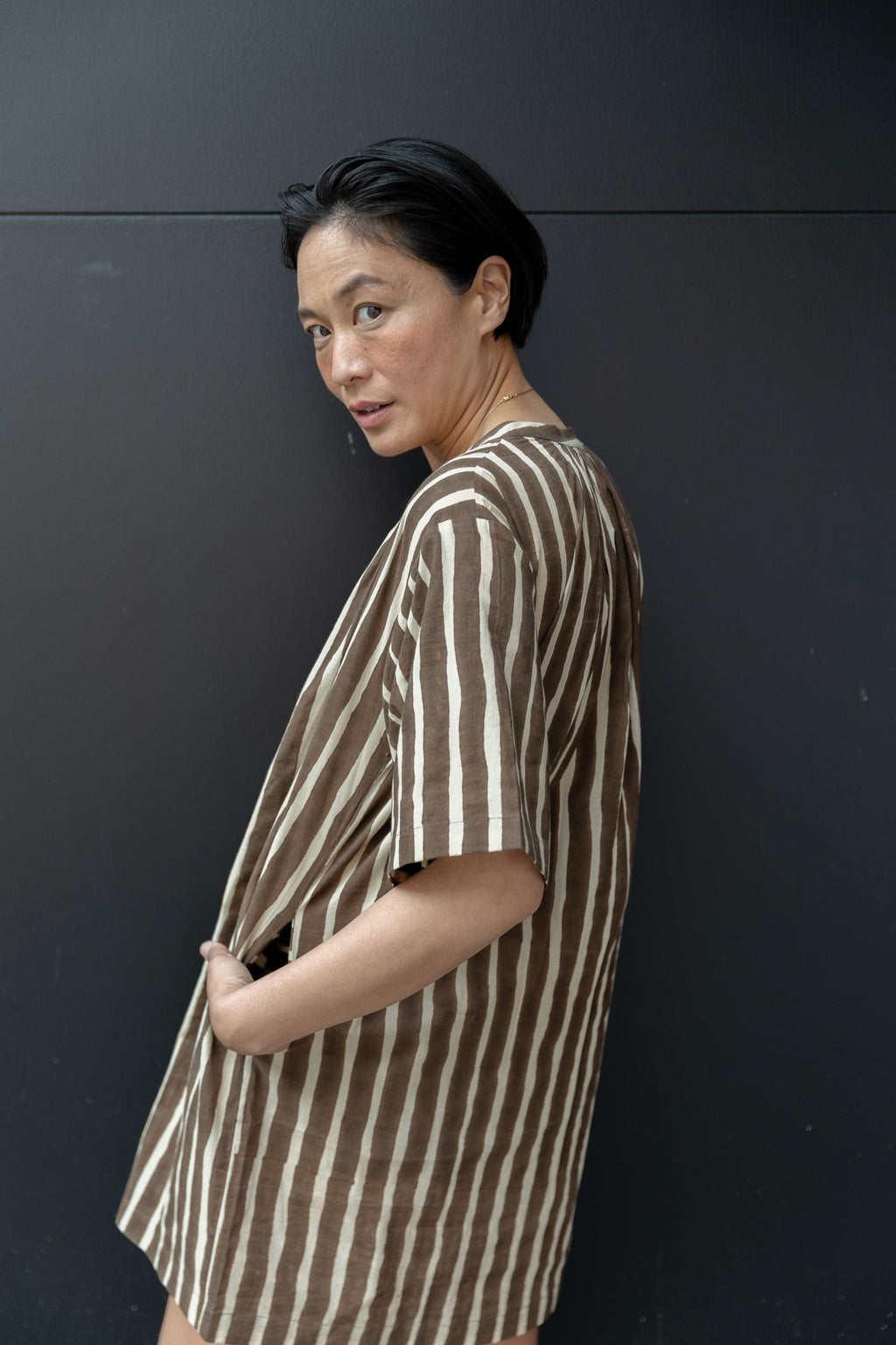 Brown and white striped tunic-style shirt with a relaxed, oversized fit. Features a round neckline with a small V-slit opening, short wide sleeves, and a slightly textured fabric. The vertical stripes create a flattering, elongated effect, and the lightweight material enhances its breezy, casual appeal.