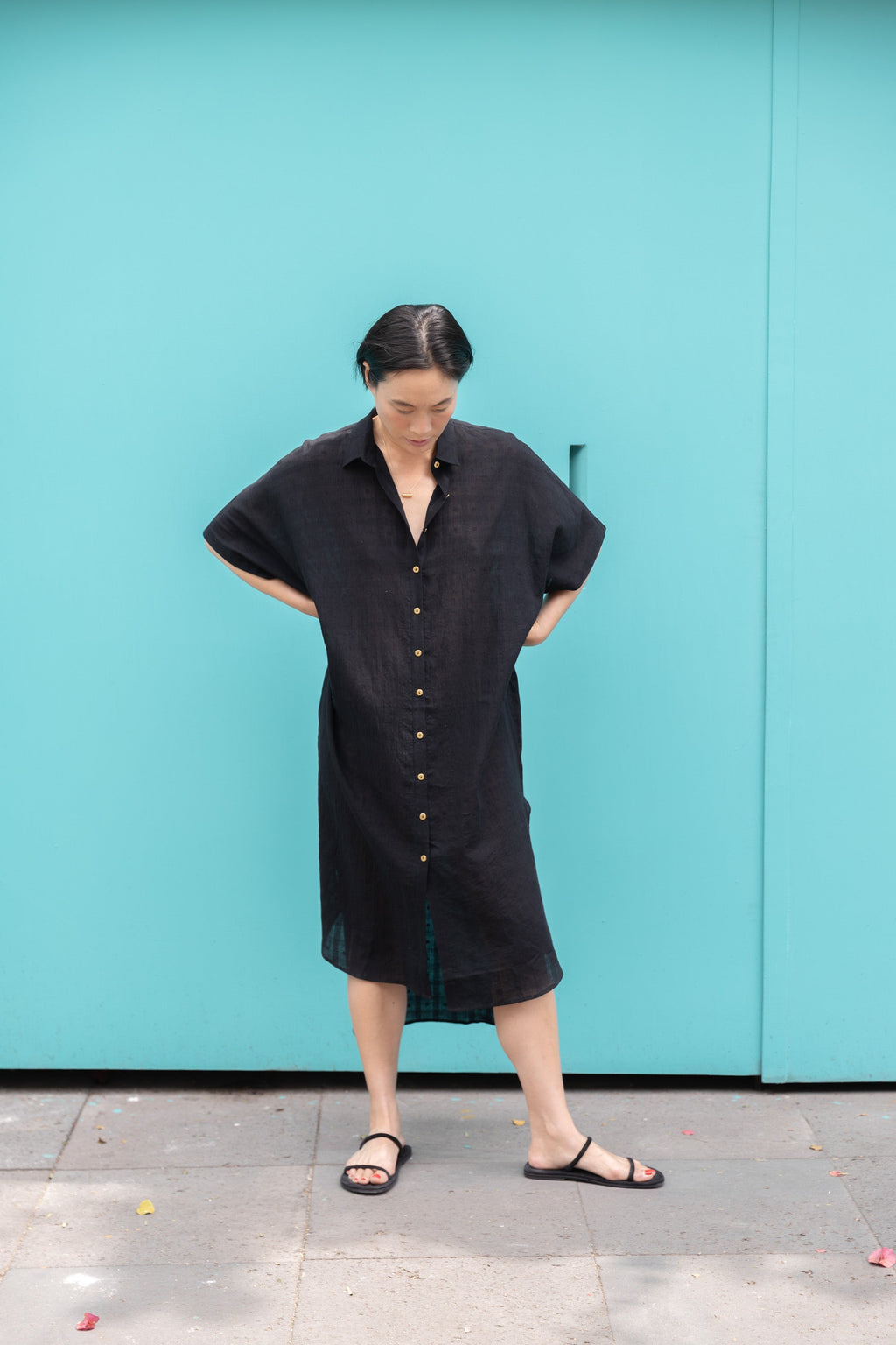 Black button-down shirt dress crafted from breathable, textured linen fabric. Designed with a relaxed, oversized silhouette for effortless movement, featuring a classic collar and short sleeves. The curved high-low hemline adds a modern touch, while the row of contrasting buttons enhances the structure. A versatile, airy piece perfect for everyday wear.