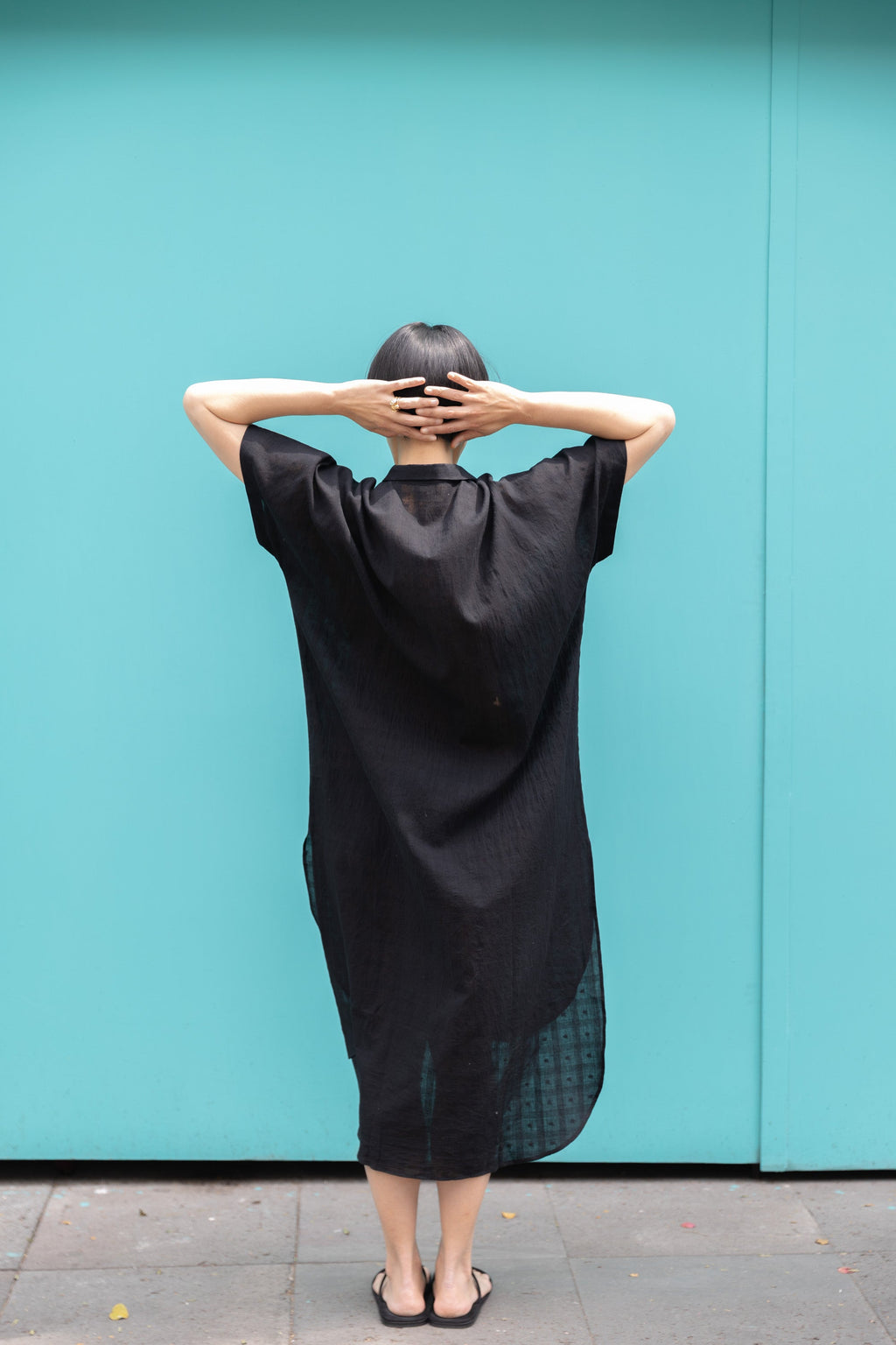 Black button-down shirt dress crafted from breathable, textured linen fabric. Designed with a relaxed, oversized silhouette for effortless movement, featuring a classic collar and short sleeves. The curved high-low hemline adds a modern touch, while the row of contrasting buttons enhances the structure. A versatile, airy piece perfect for everyday wear.