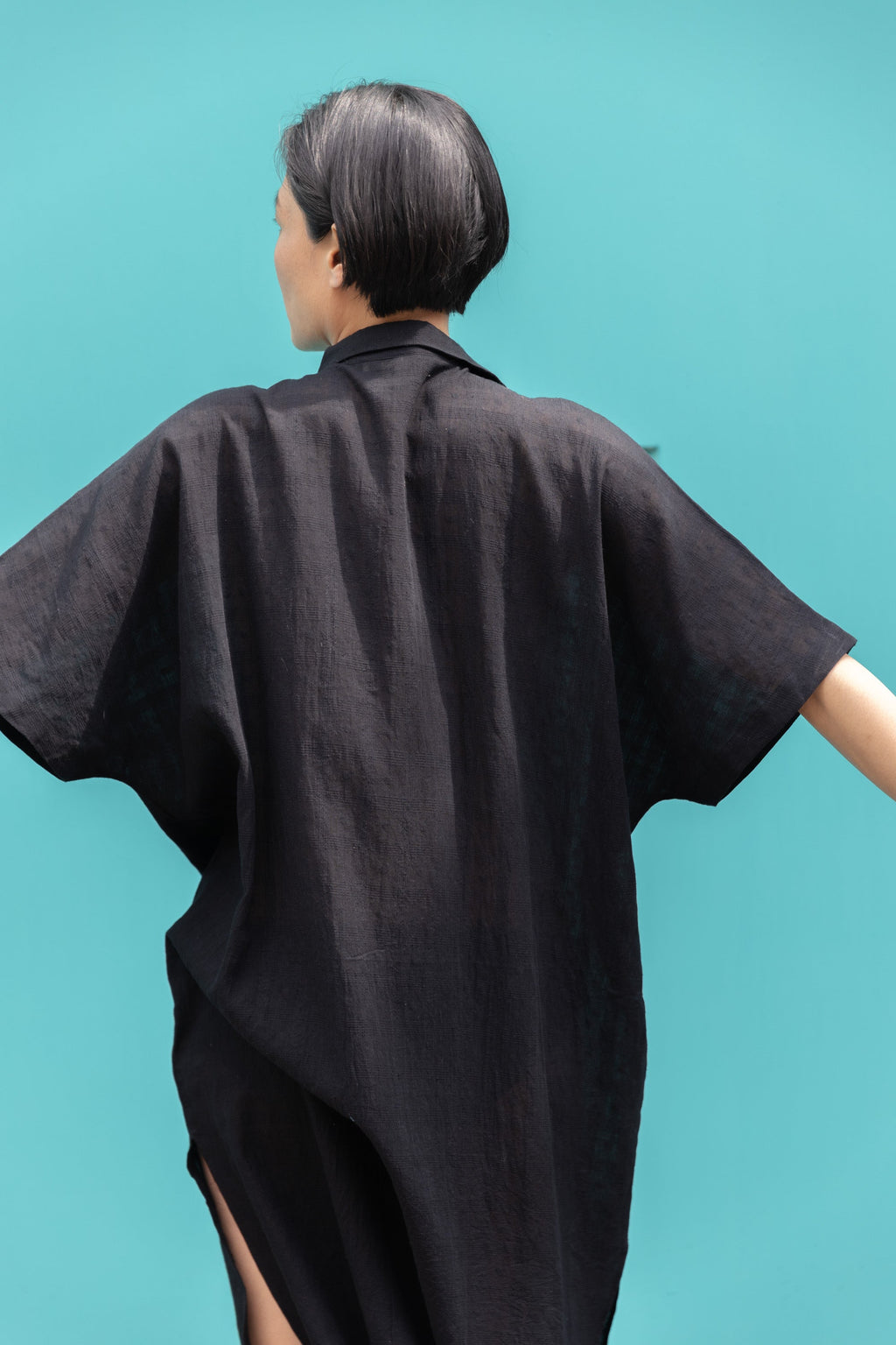 Black button-down shirt dress crafted from breathable, textured linen fabric. Designed with a relaxed, oversized silhouette for effortless movement, featuring a classic collar and short sleeves. The curved high-low hemline adds a modern touch, while the row of contrasting buttons enhances the structure. A versatile, airy piece perfect for everyday wear.