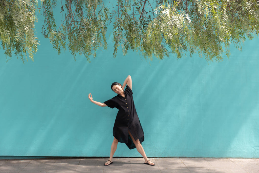 Black button-down shirt dress crafted from breathable, textured linen fabric. Designed with a relaxed, oversized silhouette for effortless movement, featuring a classic collar and short sleeves. The curved high-low hemline adds a modern touch, while the row of contrasting buttons enhances the structure. A versatile, airy piece perfect for everyday wear.