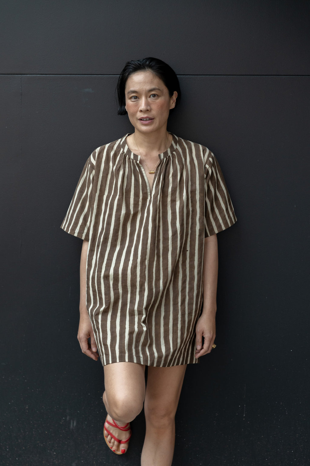 Brown and white striped tunic-style shirt with a relaxed, oversized fit. Features a round neckline with a small V-slit opening, short wide sleeves, and a slightly textured fabric. The vertical stripes create a flattering, elongated effect, and the lightweight material enhances its breezy, casual appeal.