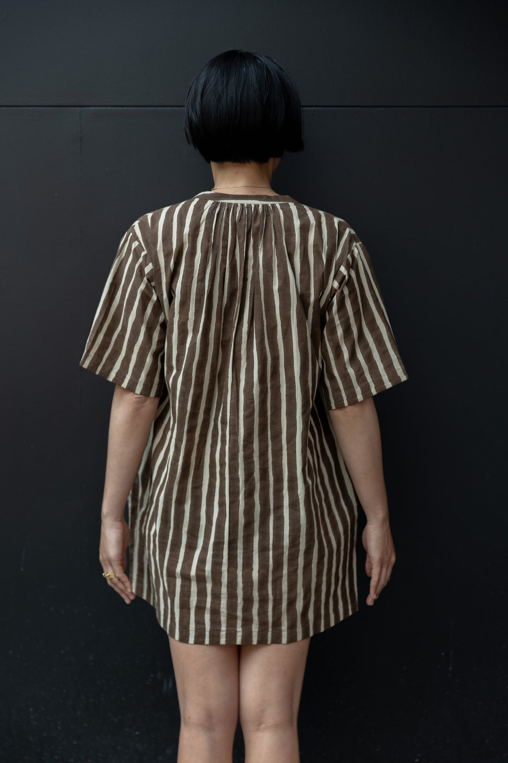 Brown and white striped tunic-style shirt with a relaxed, oversized fit. Features a round neckline with a small V-slit opening, short wide sleeves, and a slightly textured fabric. The vertical stripes create a flattering, elongated effect, and the lightweight material enhances its breezy, casual appeal.